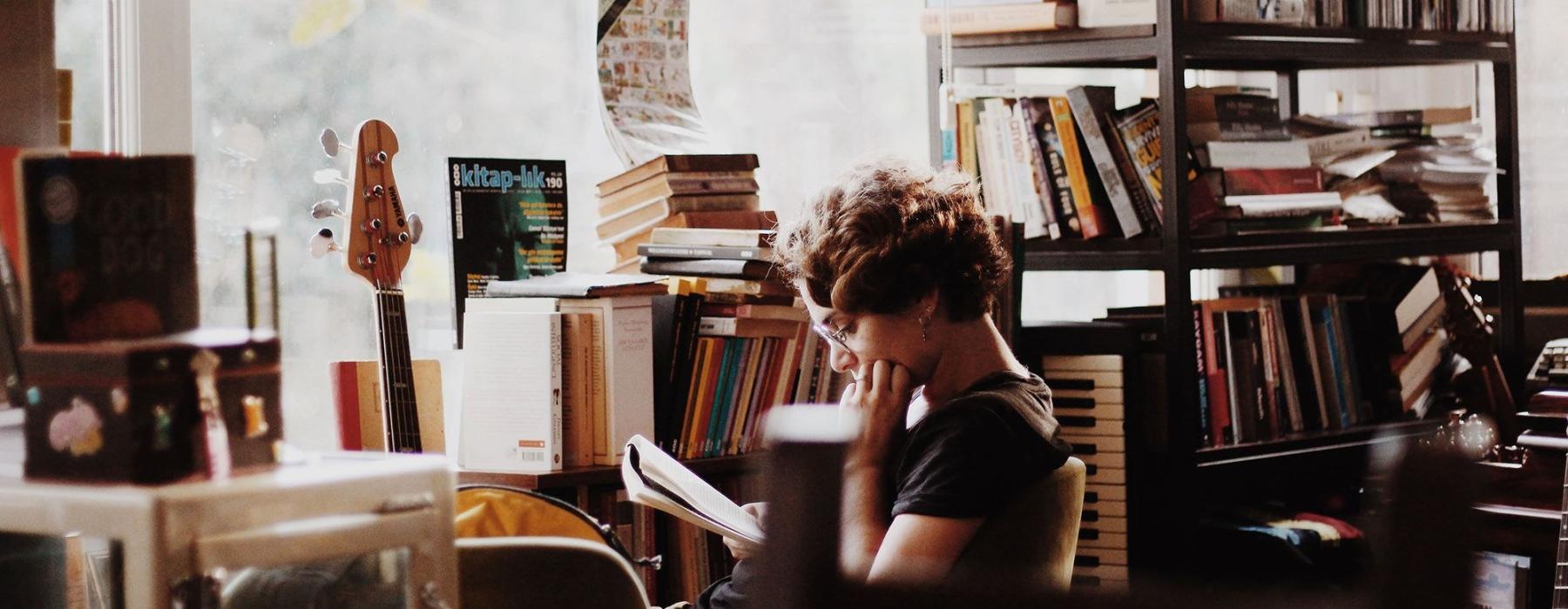 woman, surrounded by books, sits and reads in front of a picture window